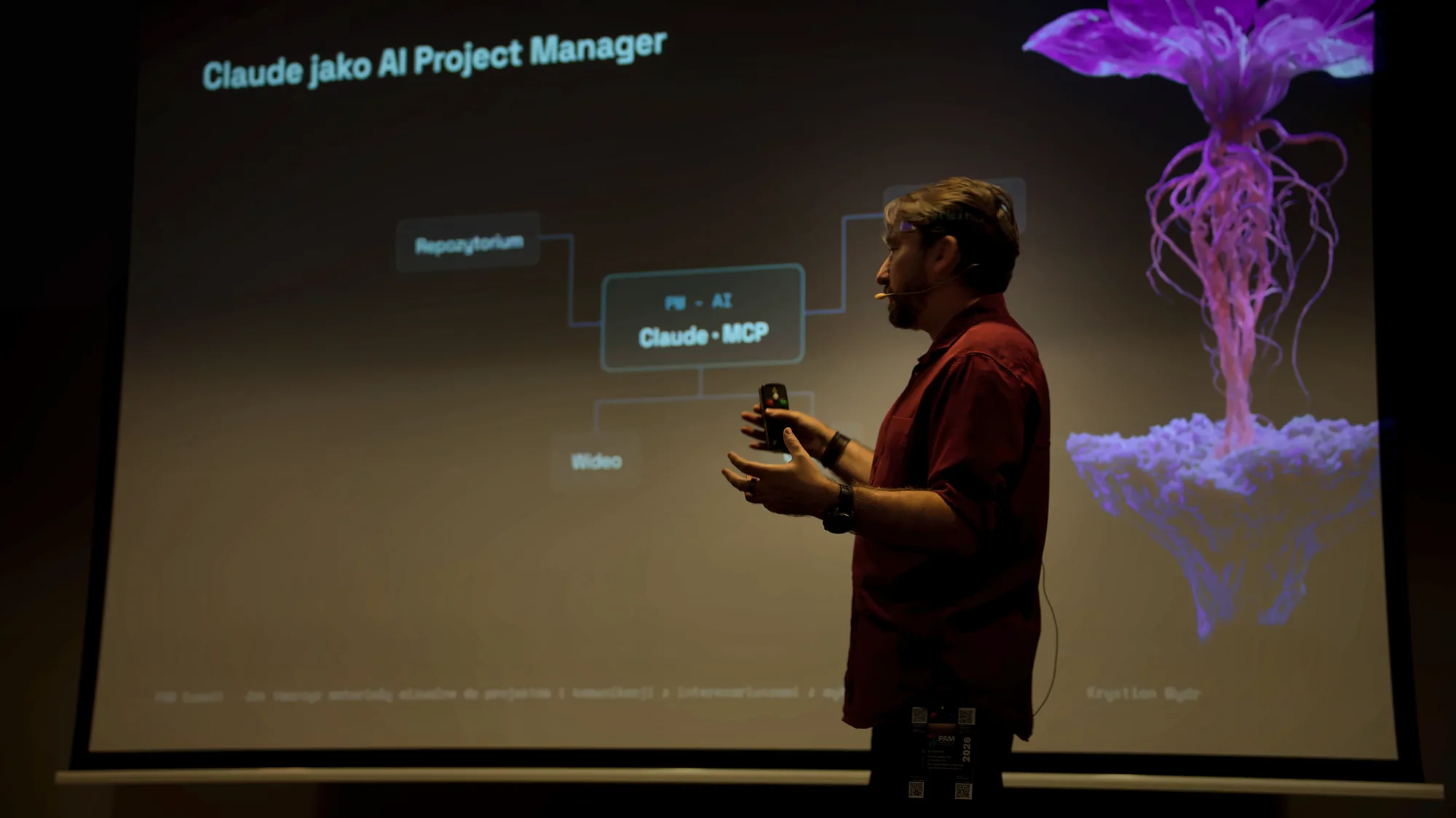 Krystian Wydro w bordowej koszuli z pilotem w dłoni na scenie PAM Summit 2026 w Krakowie, za nim slajd „Claude jako AI Project Manager” ze schematem Claude i MCP