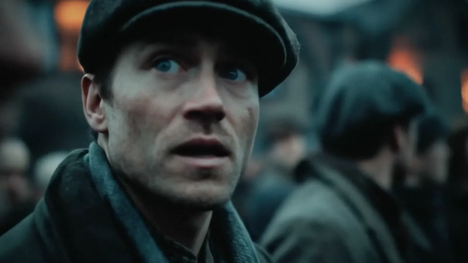 Close-up of a man in a flat cap, flames behind him