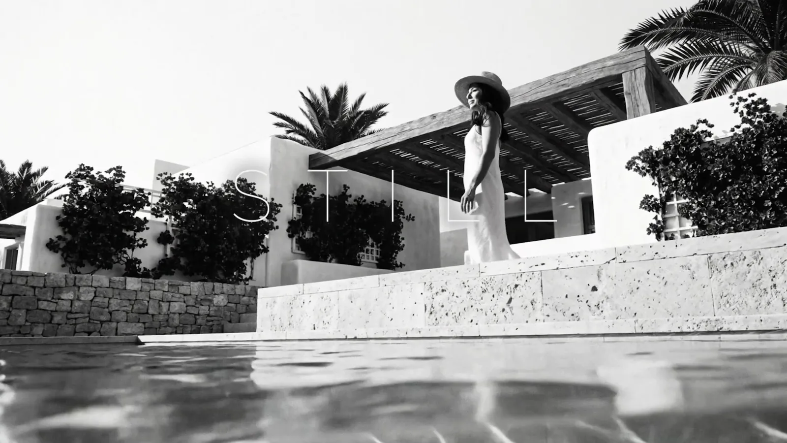 A woman in a hat on a terrace above the pool of a white house