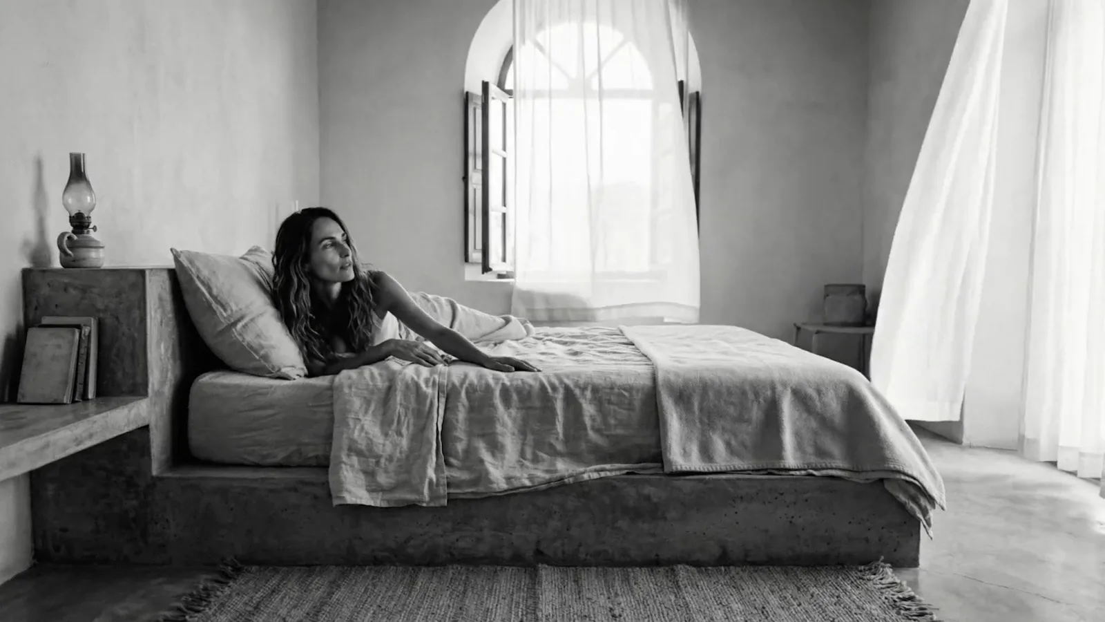 A woman resting on a bed by a window with a curtain